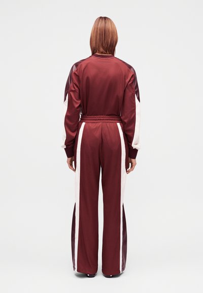 Bordó poliészter melegítő fehér oldalsávokkal. Laza fazon, rugalmas derékpánt és hosszú ujjú, kontrasztos vállrészletekkel.