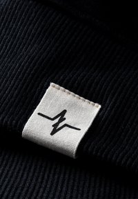 Zwarte ribstof met een beige label met een zwart bliksemschichtlogo, stevig aan de rand gestikt.