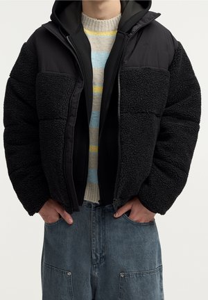 Personne portant une veste polaire noire et matelassée par-dessus un pull rayé, mains dans les poches d'un pantalon en jean bleu.