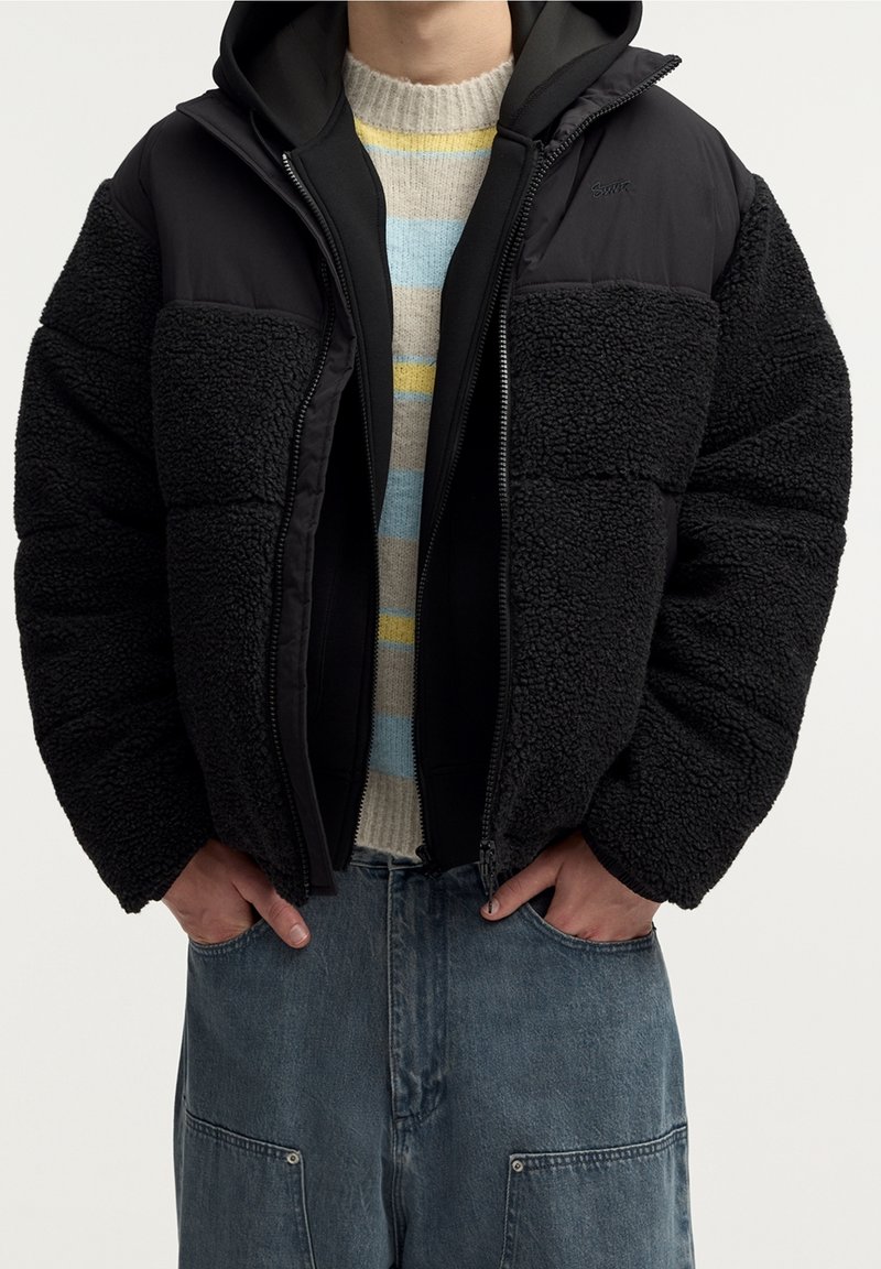Personne portant une veste polaire noire et matelassée par-dessus un pull rayé, mains dans les poches d'un pantalon en jean bleu.