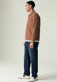 Camisa marrón de manga larga con botones, llevada sobre una camiseta blanca. Combinada con unos jeans azul oscuro y zapatillas blancas con acentos negros.