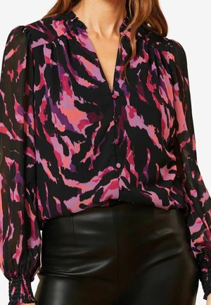 Femme portant un chemisier à manches longues avec un imprimé abstrait violet, rose et noir, associé à un pantalon en cuir noir taille haute.