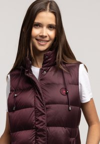 Burgundowy kamizelka puchowa z wysokim kołnierzem, zamkiem błyskawicznym z przodu i sznurkiem do regulacji. Posiada naszywkę z logo oraz błyszczącą teksturę materiału. Nosi się na białej koszuli.