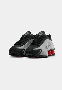 Um par de sapatilhas de corrida Nike Shox, em preto e cinza, com colunas vermelhas de absorção de choque e atacadores pretos, sobre um fundo branco.