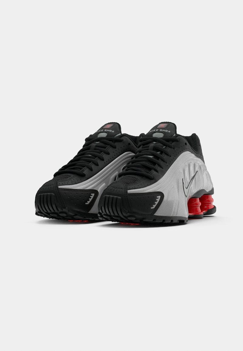 Une paire de chaussures de course Nike Shox noires et grises avec des colonnes d'amortissement rouges et des lacets noirs sur un fond blanc.