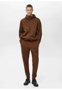 Felpa con zip marrone, colletto alto e tasche anteriori, abbinata a pantaloni marroni coordinati. Presenta una texture liscia e morbida e un design essenziale.