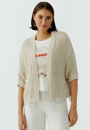 Jonge vrouw die een beige open-geweven vest, een wit T-shirt met een afbeelding, lichte broek en oorringen draagt, staand tegen een effen achtergrond.