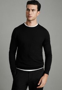 Pulover negru din tricot, cu guler rotund, mâneci lungi și manșete din bumbac, purtat peste o cămașă albă stratificată. Textură netedă și design ajustat.