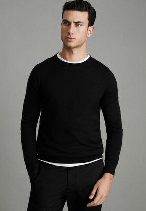 Pull en maille noire avec col rond, manches longues et poignets côtelés, porté par-dessus une chemise blanche à couches. Texture lisse et design ajusté.