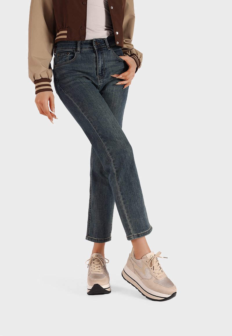 Sneakers eleganti in beige metallico con tessuto texturizzato, abbinati a jeans blu scuro a gamba dritta e a una giacca bomber marrone.