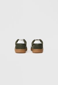 Ein Paar dunkelgrüne Wildleder-Sneaker mit weißen Fersenakzenten und hellbraunen Gummisohlen, von hinten auf einem schlichten weißen Hintergrund betrachtet.