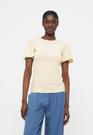 Γυναίκα όρθια, φοράει μπλούζα κρεμ με κοντά μανίκια και φαρδιά μπλε παντελόνια, μπροστά σε απλό λευκό φόντο.