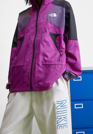 Person iført en lilla og sort North Face jakke med lynlås foran og hvide Nike joggingbukser med blå bogstaver.