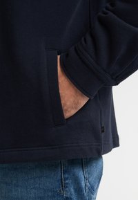 Donkerblauwe sweatshirt met een zichtbare zijzak, geribbelde mouwen en een zachte stofstructuur. Gedragen over lichtblauwe jeans.