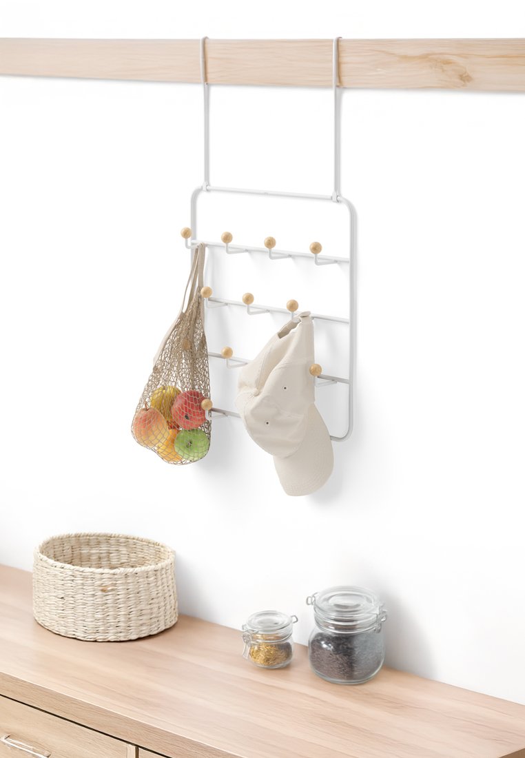 Biały metalowy regał na ścianę z drewnianymi hakami, trzymający beżową czapkę i siatkową torbę z jabłkami, zamontowany na jasnej drewnianej powierzchni.
