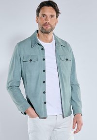 Hellgrüne Wildlederjacke mit Kragen, durchgehender Knopfleiste und zwei Brusttaschen. Verfügt über schwarze Knöpfe und eine glatte Textur.