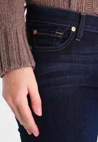 Jeans de ganga azul-escuro com costura dourada, apresentando bolsos frontais e um suéter castanho acanelado. Foco na textura e na estrutura do tecido.