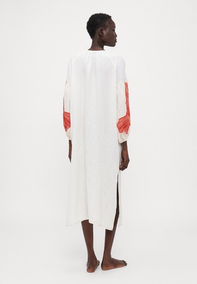 Weißes Leinenkleid mit langen, gepufften Ärmeln und orange-roten Akzenten. Es hat einen langen, geraden Schnitt mit seitlichen Schlitzen für mehr Bewegungsfreiheit.