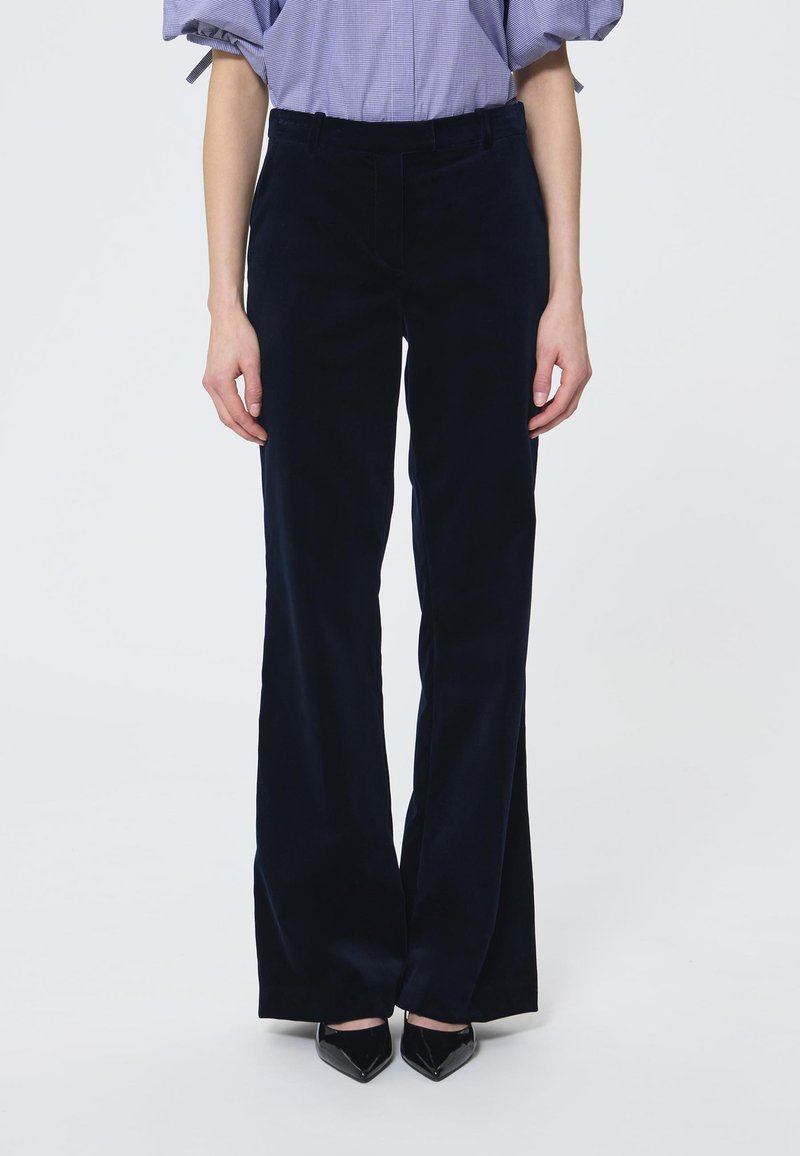 Tamsiai mėlynos veliūro kelnės su platėjančiomis kojomis, pasižyminčios lygiu paviršiumi ir standartiniu liemeniu. Dėvimos su smailiais juodais aukštakulniais.