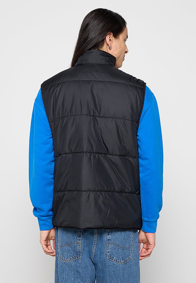 Schwarze gepolsterte Weste mit hohem Kragen, genähten Paneelen und glatter Textur, getragen über einem blauen Langarmshirt.
