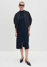 Navy blue midi dress with a front slit, paired with a black bomber jacket. Footwear features pointed toes and a slingback design.
