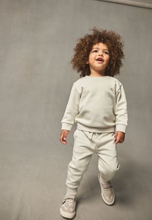 Lyse beige sweatshirt og matchende sweatpants, begge med en blød tekstur. Accentlogo på brystet. Barn iført lyse sneakers.