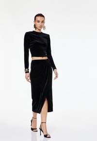 Sort fløjl langærmet crop top parret med en knælang nederdel med en sideslids. Accentueret med guldsmykker og strophæle.