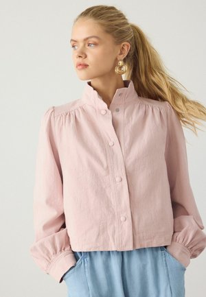 Ung kvinde med blond hestehale iført guldringearreringe, en lys pink jakke med knapper og lyseblå bukser, der kigger mod venstre mod en ensfarvet baggrund.