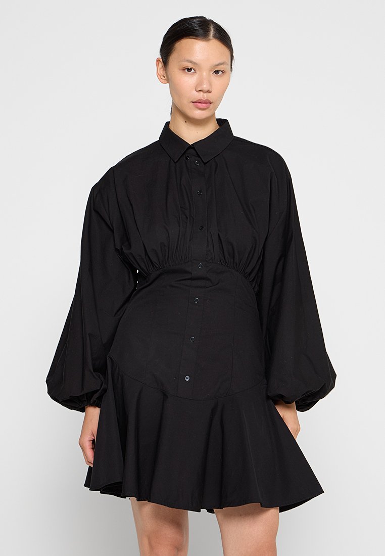 Eine Frau trägt ein schwarzes Hemdkleid mit langen Puffärmeln, Knopfleiste, taillierter Taille und ausgestelltem Rock vor einem weißen Hintergrund.