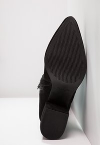 Musta sametista valmistettu korkokenkä, jossa on terävä kärki, teksturoitu pohja ja koristeellinen vetskari kyljessä. Sujuva viimeistely hienovaraisella ompelulla.