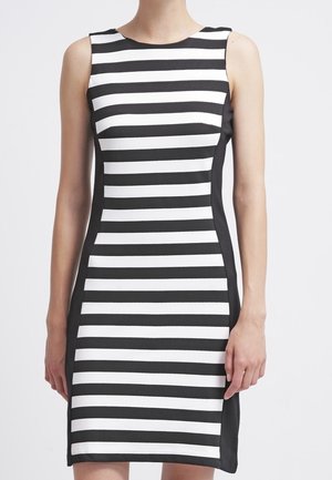 Sukienka bez rękawów do kolan z czarno-białymi poziomymi paskami i jednolitymi czarnymi panelami po bokach, noszona przez stojącą osobę.