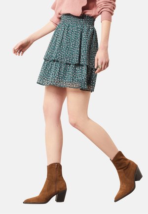 Femme portant une mini-jupe verte fleurie à plusieurs couches, un pull rose et des bottines marron en suède à talons, marchant vers l'avant.