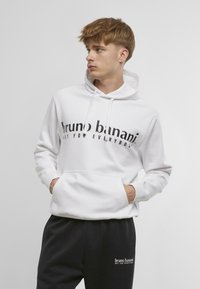 Fehér kapucnis póló fekete felirattal: "bruno banani NEM MINDENKINEK," kanguru zsebbel és állítható zsinórral a kapucniban.