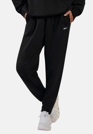 Persona con pantalones deportivos negros de Reebok con las manos en los bolsillos y zapatillas blancas para correr, de pie con las piernas cruzadas.