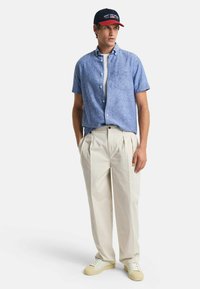 Blauw short-sleeve overhemd met knoopsluiting, beige geplooide broek, wit t-shirt eronder, marineblauwe pet met rode accenten en lichte sneakers.