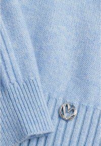 Sanftes hellblaues Strickgewebe mit gerippten Kanten und einem kleinen silbernen herzförmigen Metallschmuck, der nahe dem unteren Rand angebracht ist.