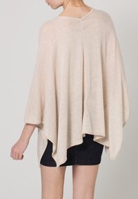 Donna che indossa un poncho lavorato a maglia beige, morbido e con una texture a coste, su pantaloni corti neri, vista da dietro su uno sfondo neutro.