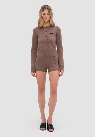 Femme debout, portant un cardigan marron boutonné et un short assorti avec des chaussures noires ouvertes aux orteils, style slip-on, sur fond clair uni.