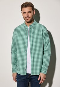 Green plaid button-up shirt with a collar, long sleeves, and a front pocket, worn over a white t-shirt, paired with blue jeans.