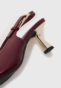 Burgundijas lakota ādas slingback zābaks ar zeltītu ģeometrisku sprādzi un atstarojošu smilšu pulksteņa formas zelta papēdi uz baltā fona.
