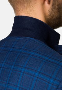 Niebieska koszula w kratę o teksturowanej tkaninie, z ciemnym kołnierzykiem i kontrastującymi jaśniejszymi niebieskimi liniami w wzorze. Widok z tyłu.