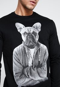 Musta pitkähihainen paita, jossa on suuri grafiikka ranskanbulldogista, joka pukeutunut harmaaseen huppariin, jossa on valkoiset kiristysnauhat ja helminauha.