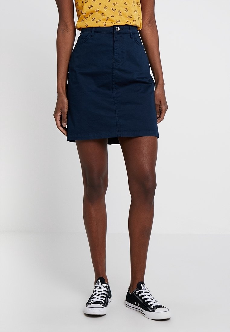 Jupe en coton bleu marine à coupe droite, avec fermeture à boutons sur le devant et deux poches latérales, associée à des baskets noires et un haut à motifs jaunes.