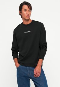 Mand iført sort langærmet Calvin Klein skjorte og blå jeans, står med den ene hånd i lommen mod en simpel hvid baggrund.