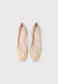 Beige leather flats with a smooth texture, almond-shaped toe, padded insole, and a small gold logo accent on the side.