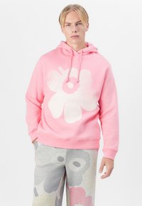 Rosa Kapuzenpullover aus weichem Material mit einem großen weißen Blumendesign. Kombiniert mit grauen Jogginghosen, die ein Blumenmuster in sanften Farben zeigen.