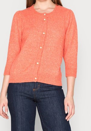 Kobieta w koralowym dzianinowym kardiganie z perłowymi guzikami i rękawami do połowy przedramienia, w połączeniu z ciemnoniebieskimi dżinsami.