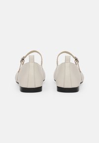 Zapatos de cuero color beige con diseño de correas en T y un tacón negro bajo; textura suave con un sutil detalle de costura en la parte trasera.