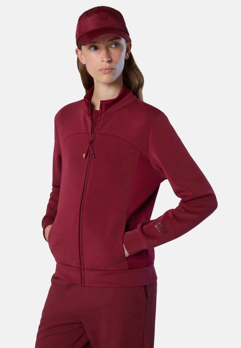 Bordeauxfarbene Zip-Jacke mit hohem Kragen, langen Ärmeln und Seitentaschen. Glatte Textur; kleines Logo-Detail am Arm.