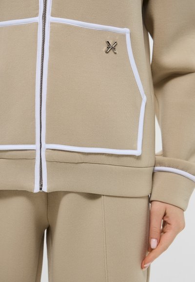 Giacca beige con zip e piping bianco, dotata di due grandi tasche frontali e un piccolo logo argentato sul petto. Tessuto morbido e testurizzato.